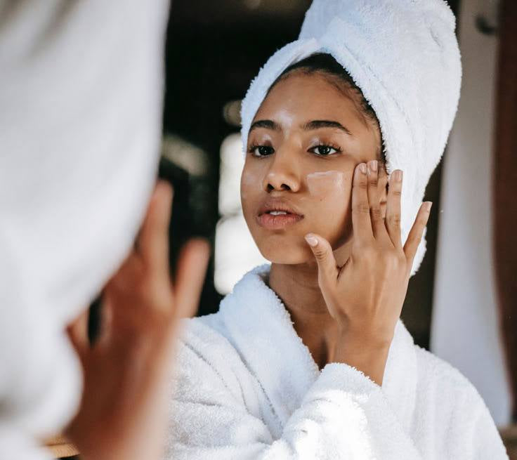 Woman in a white robe applying cream to her face with a towel on her head.